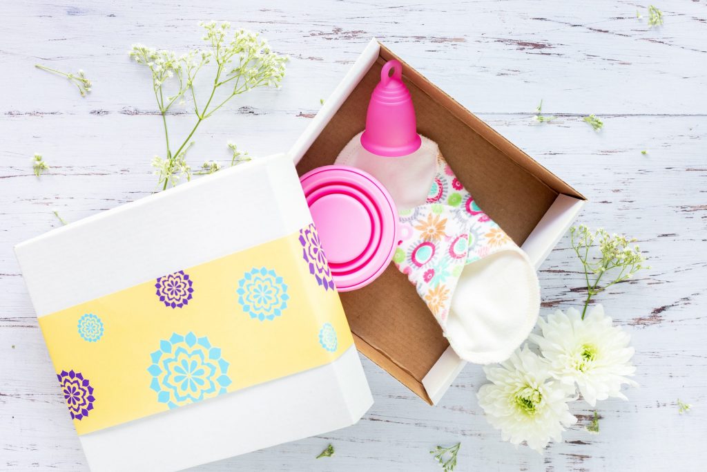 A white and yellow box sits on a white table, containing a pink mentrual cup, a reusable sanitary pad and a small pink pot